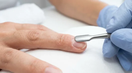 Nail tech pushing back cuticles on a bare natural nail with a metal cuticle pusher under bright salon light