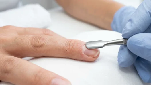 Nail tech pushing back cuticles on a bare natural nail with a metal cuticle pusher under bright salon light