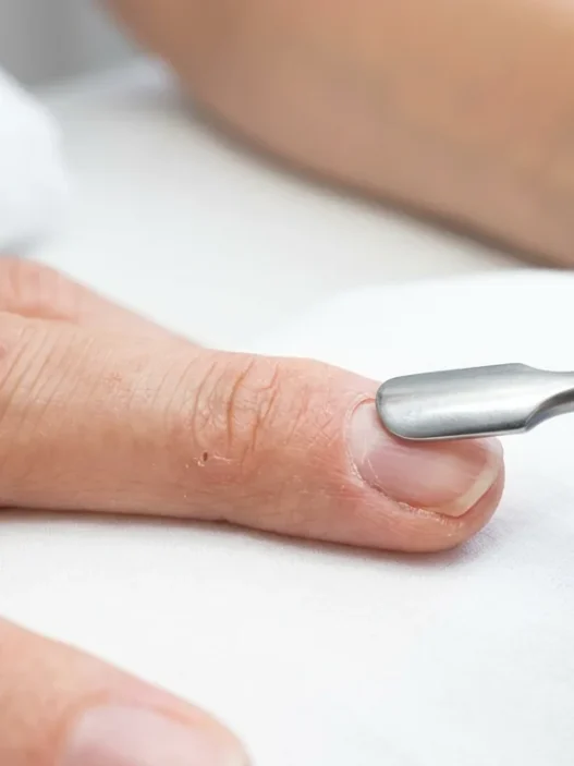 Nail tech pushing back cuticles on a bare natural nail with a metal cuticle pusher under bright salon light