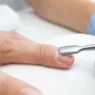 Nail tech pushing back cuticles on a bare natural nail with a metal cuticle pusher under bright salon light