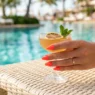 Woman holding citrus cocktail with neon coral gel nails on a sunlit pool deck with turquoise water bokeh