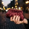 Manicured hand with deep burgundy square nails holding a satin clutch under warm amber street-lamp light at night