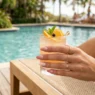 Manicured hand with dusty rose to cream ombre nails holding cocktail glass on sunlit pool deck
