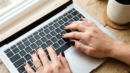 Close-up overhead of hands with short square espresso nails typing on laptop keyboard with coffee mug visible