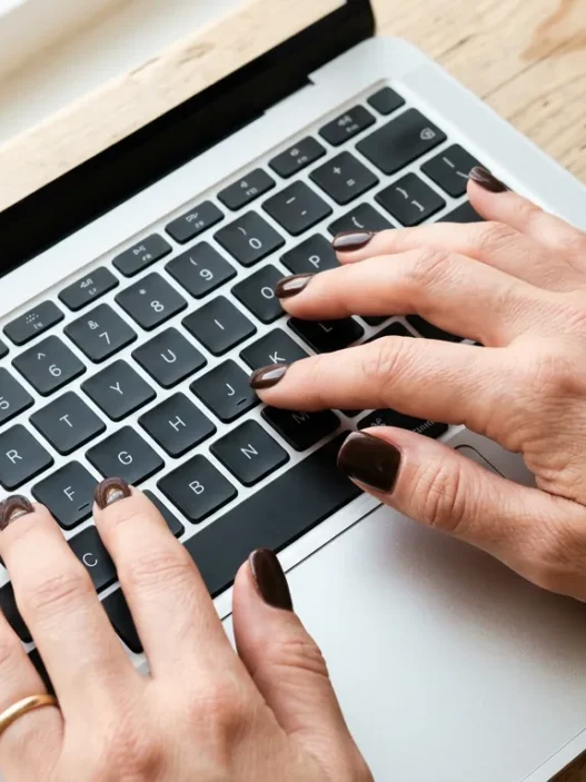 Close-up overhead of hands with short square espresso nails typing on laptop keyboard with coffee mug visible