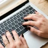 Close-up overhead of hands with short square espresso nails typing on laptop keyboard with coffee mug visible