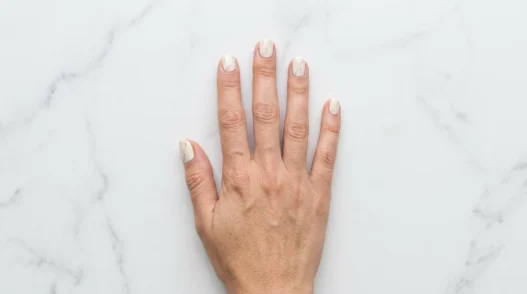 Overhead shot of hand on white marble surface showing soft cream marble nail design with delicate grey veining in daylight