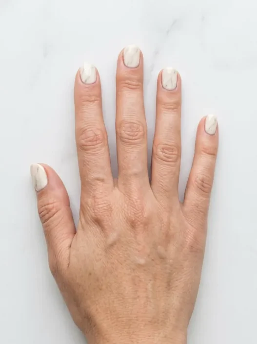 Overhead shot of hand on white marble surface showing soft cream marble nail design with delicate grey veining in daylight