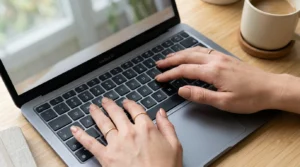 Close-up overhead of manicured hands with squoval nude nails typing on a laptop keyboard in soft window light