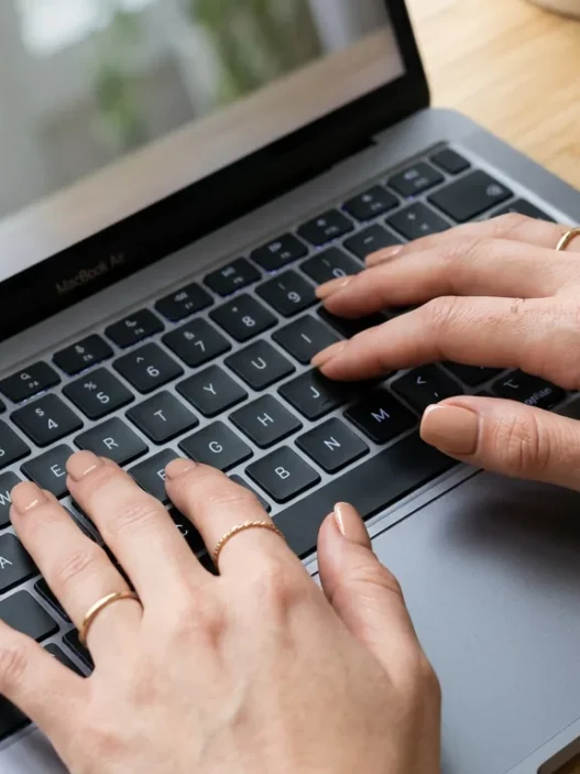 Close-up overhead of manicured hands with squoval nude nails typing on a laptop keyboard in soft window light
