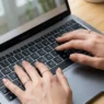Close-up overhead of manicured hands with squoval nude nails typing on a laptop keyboard in soft window light