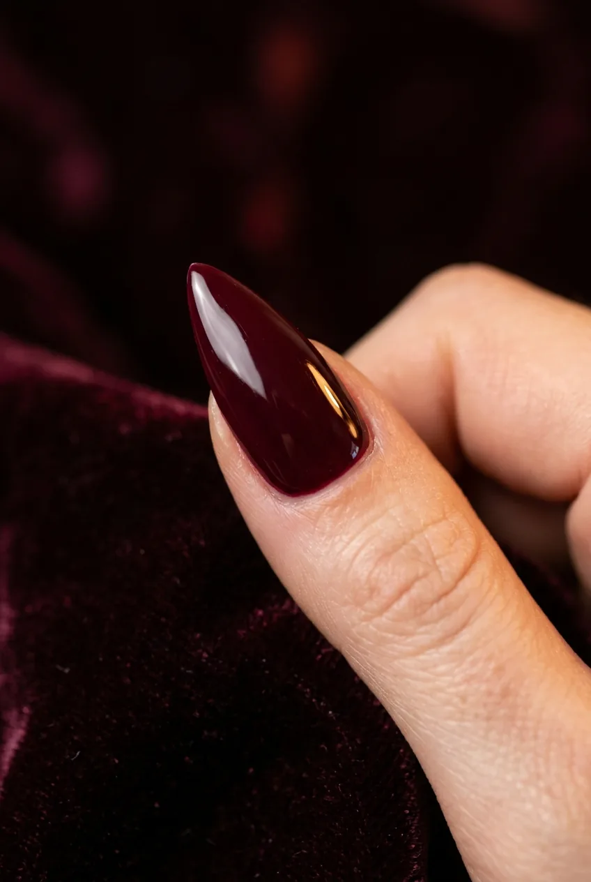 Extreme close-up of a single stiletto nail in glossy burgundy showing curved apex structure against dark velvet background