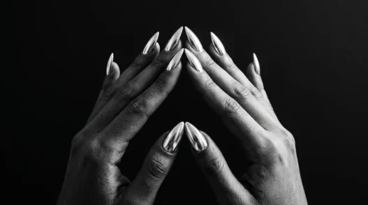 Two manicured hands with chrome silver stiletto nails pressed fingertip-to-fingertip against a matte black background under dramatic side lighting