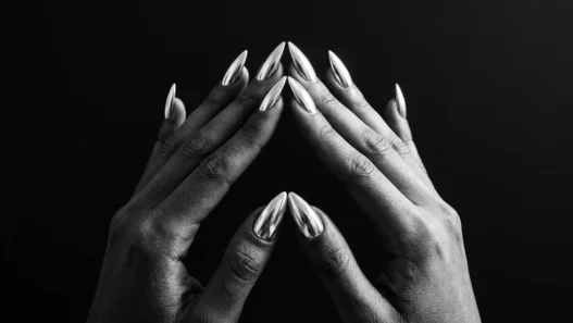 Two manicured hands with chrome silver stiletto nails pressed fingertip-to-fingertip against a matte black background under dramatic side lighting