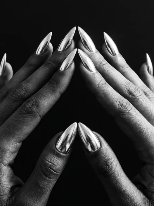 Two manicured hands with chrome silver stiletto nails pressed fingertip-to-fingertip against a matte black background under dramatic side lighting