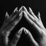 Two manicured hands with chrome silver stiletto nails pressed fingertip-to-fingertip against a matte black background under dramatic side lighting