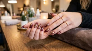 Woman's hands fanned out on salon table showing ten stiletto nails with burgundy and gold freehand designs, warm lighting
