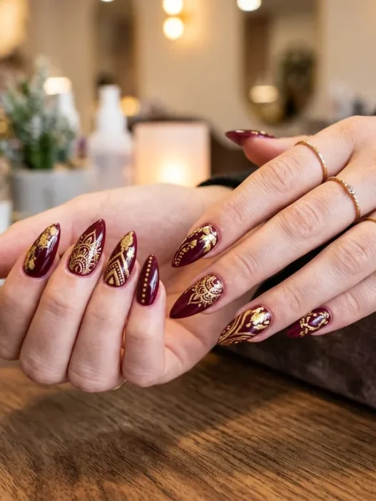 Woman's hands fanned out on salon table showing ten stiletto nails with burgundy and gold freehand designs, warm lighting