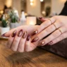 Woman's hands fanned out on salon table showing ten stiletto nails with burgundy and gold freehand designs, warm lighting