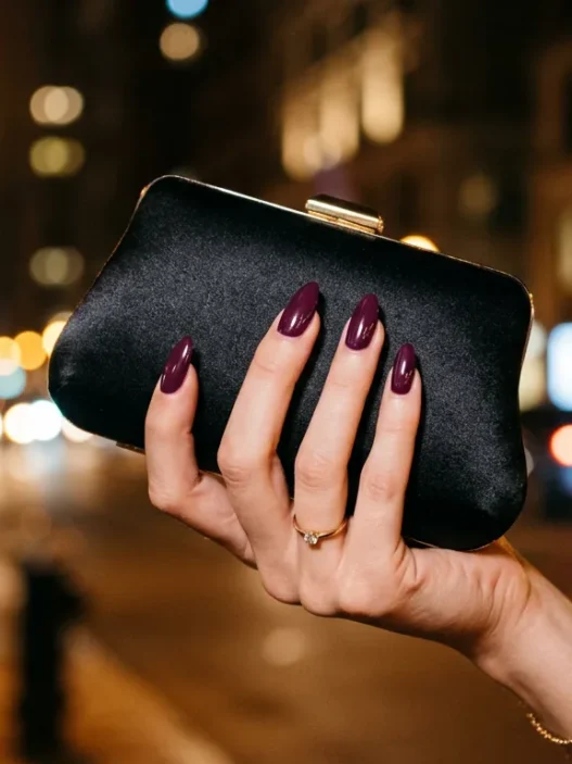 Close-up of manicured hand with deep plum stiletto nails holding black satin clutch under amber street-lamp light at night