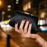 Close-up of manicured hand with deep plum stiletto nails holding black satin clutch under amber street-lamp light at night