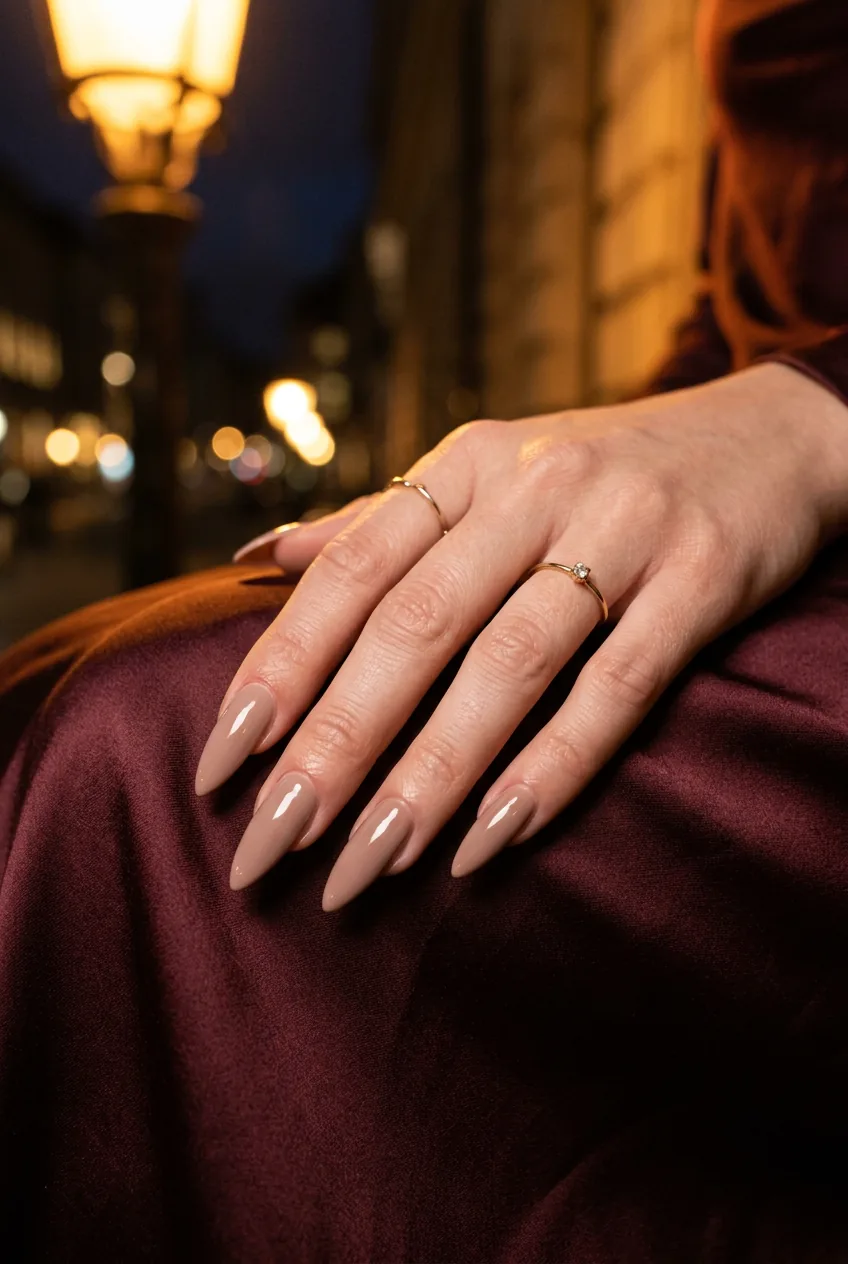 Close-up of taupe stiletto nails on a hand resting on silk fabric with healthy cuticles visible under warm amber light