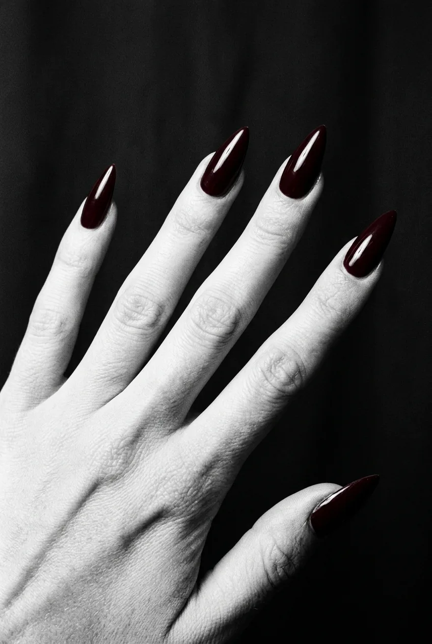 Manicured hand with wide nail beds sporting deep saturated burgundy stiletto nails fanned flat under harsh side light on matte black background